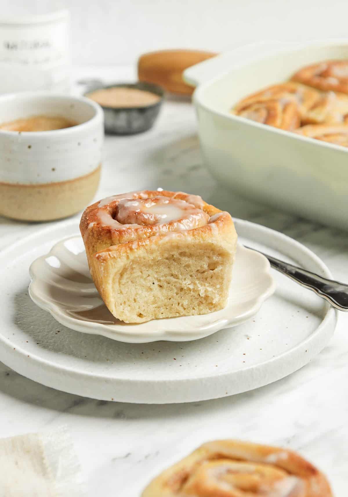 One finished cinnamon roll on a white plate with a fork and a mug of coffee in the background. 