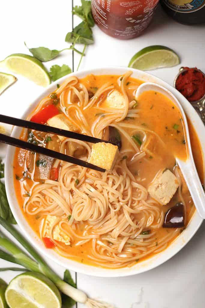 Chopsticks picking up tofu from a bowl of fresh Thai noodle soup