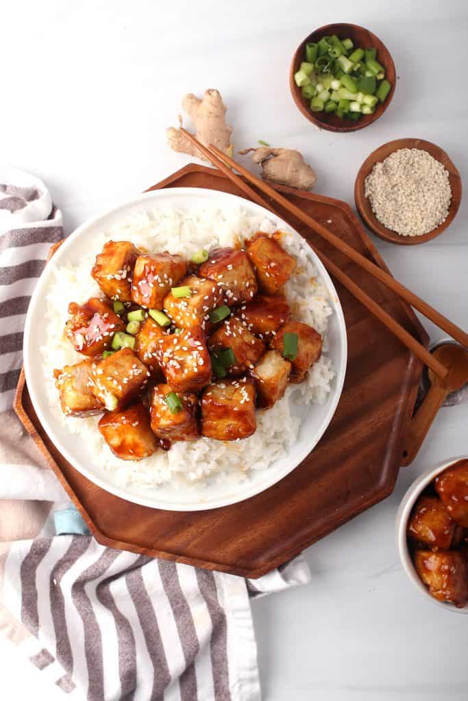 overhead shot of vegan general tso's tofu in a bowl on top of sticky rice garnished with green onions and sesame seeds