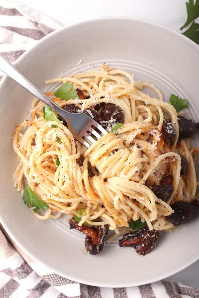 close up shot of a bite of vegan carbonara pasta twirled around a fork in a white bowl filled with pasta and shiitake bacon