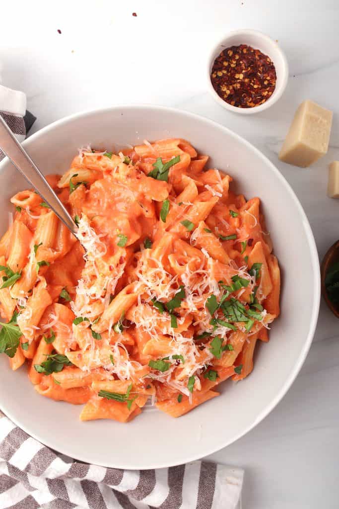 overhead shot of penne pasta tossed in vegan vodka sauce and topped with vegan parmesan and parsley in a white bowl with a silver fork