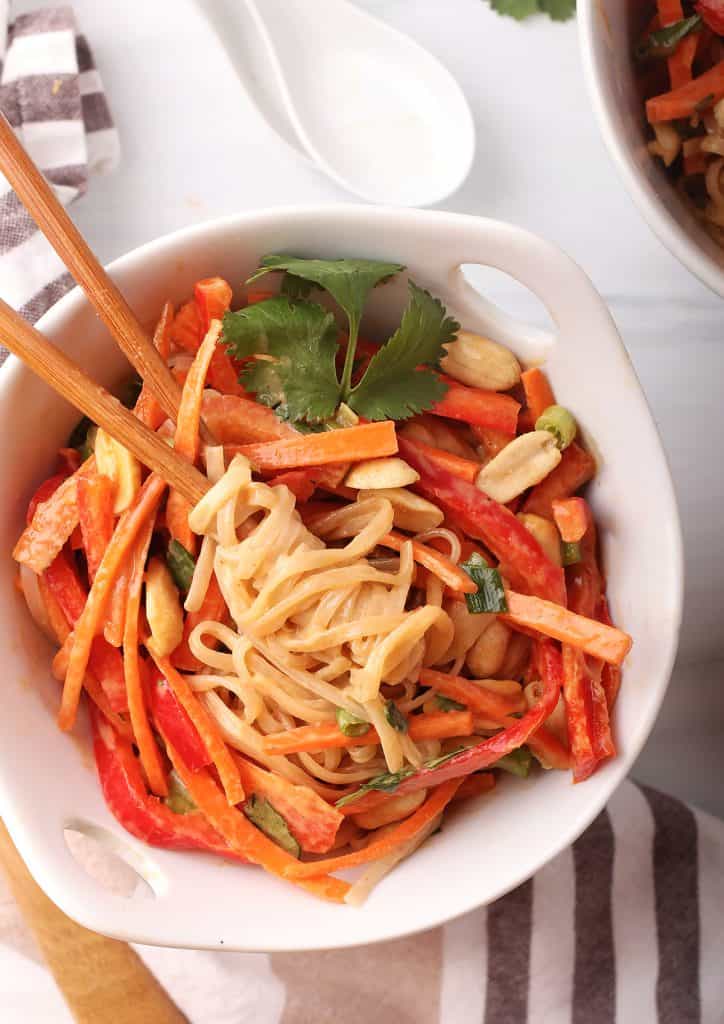 overhead close up of a single bowl of vegan thai peanut noodles in a white bowl with wooden chopsticks holding a bite