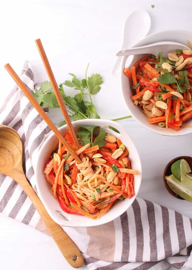 overhead shot of two white bowls filled with vegan Thai peanut noodles garnished with peanuts and cilantro on a white table with wooden chopsticks and white Asian soup spoons