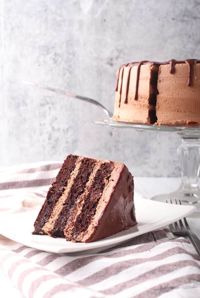 Slice of vegan chocolate cake on a white plate with the whole cake in the background. 