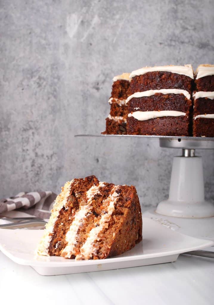 vegan carrot cake on a cake stand in the background with a slice on a white plate in the foreground