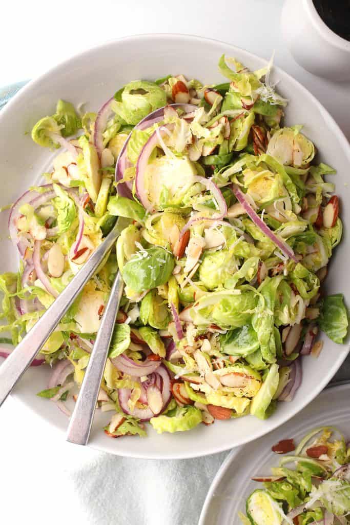 close up shot of shaved brussels sprouts salad in a white serving bowl with silver serving spoons