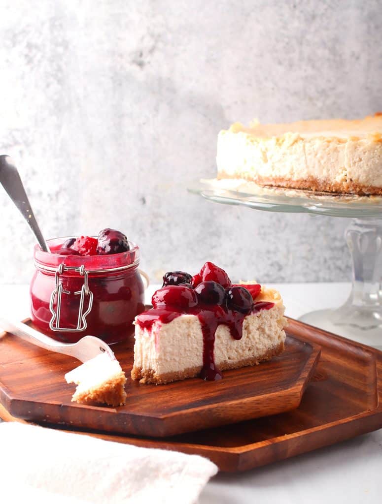 slice of new york style vegan cheesecake on an octagonal wooden plate with a silver fork and a jar of berry compote on the side