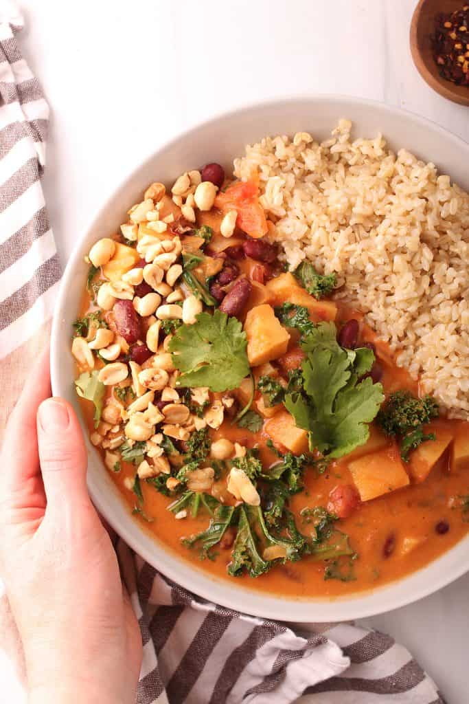 hand holding a white bowl filled with vegan peanut stew and rice