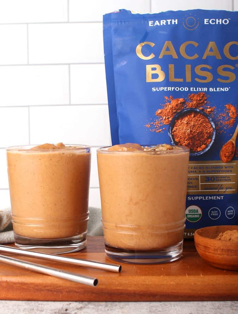 sideways shot of two clear glasses with chocolate breakfast smoothie on a wooden surface with metal straws and a bag of cacao bliss