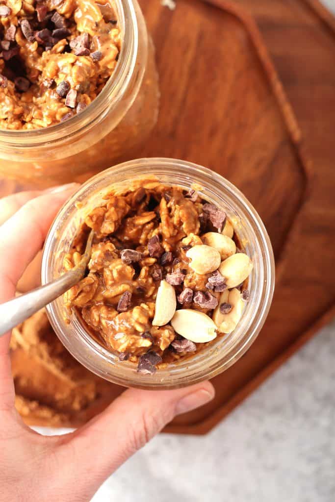overhead shot of a hand grabbing a mason jar filled with overnight oats topped with chopped peanuts and cacao nibs