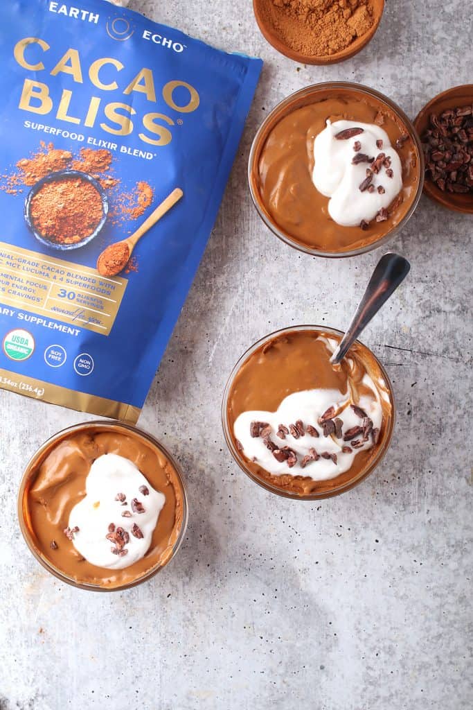 overhead shot of three cups of healthy superfood chocolate pudding on a grey table next to a bag of cacao bliss