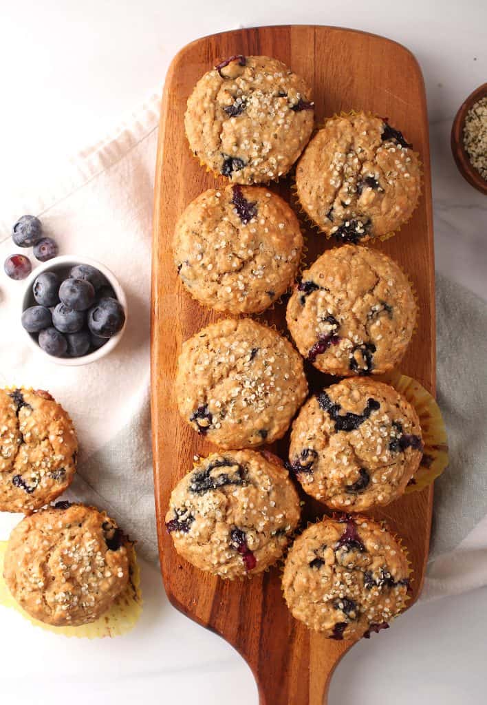 8 healthy blueberry hemp muffins on a wooden cutting board with two off to the side on a white tabletop