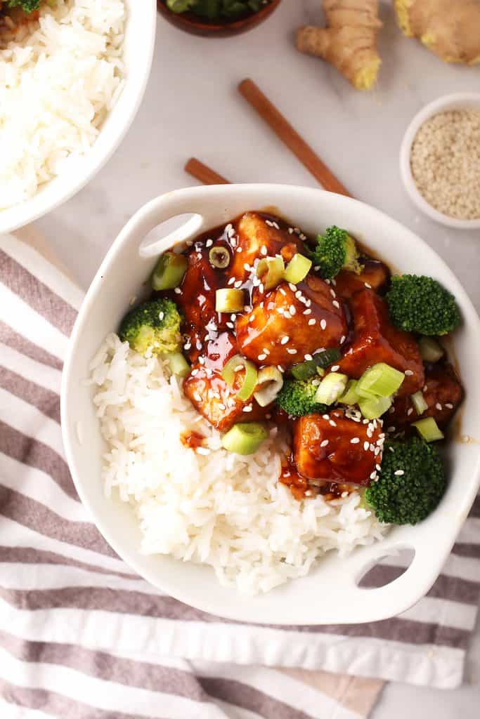 white bowl with sticky rice, teriyaki tofu and steamed broccoli with a grey and white striped towel