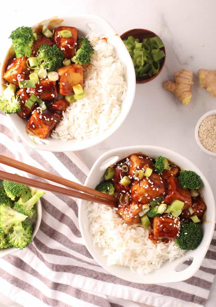 overhead shot of a white bowl with handles filled with steamed rice, teriyaki tofu and steamed broccoli with a pair of wooden chopsticks