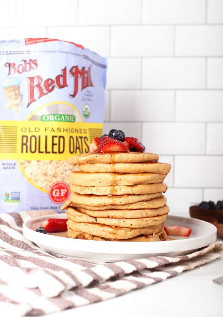 tall stack of vegan oatmeal pancakes topped with berries and maple syrup on a white plate in front of white subway tile