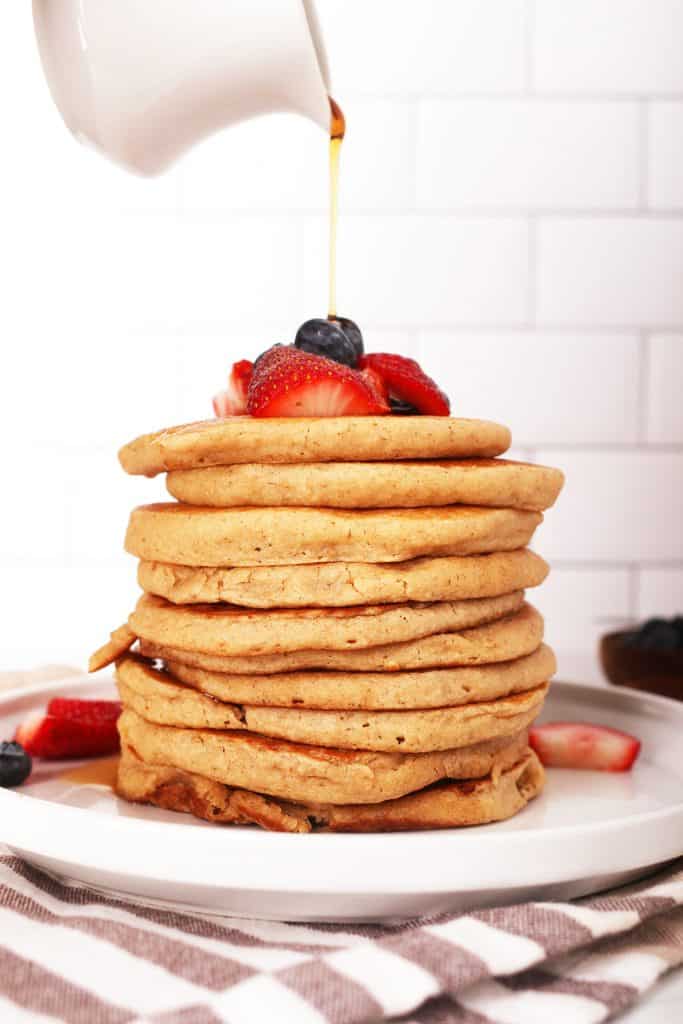 hand pouring a white carafe of syrup over a tall stack of oatmeal pancakes