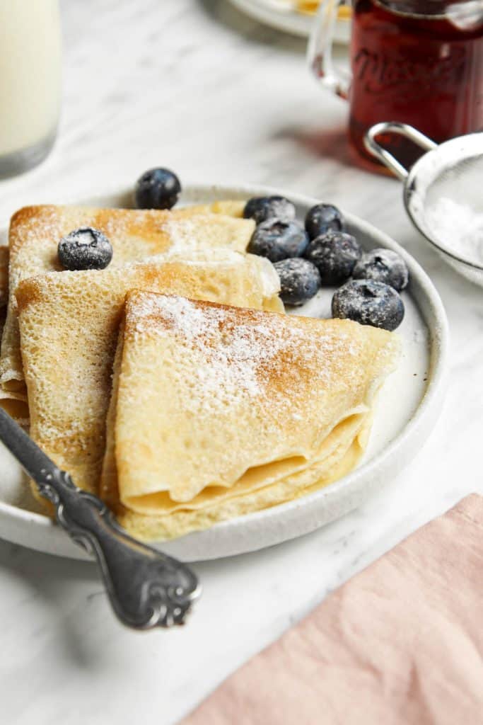 three crepes served on a white plate with blueberries, powdered sugar, a sieve and a fork.