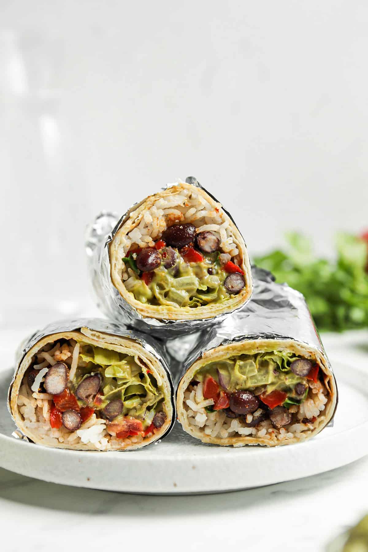 Three vegan burritos wrapped in foil and stacked on a white plate 