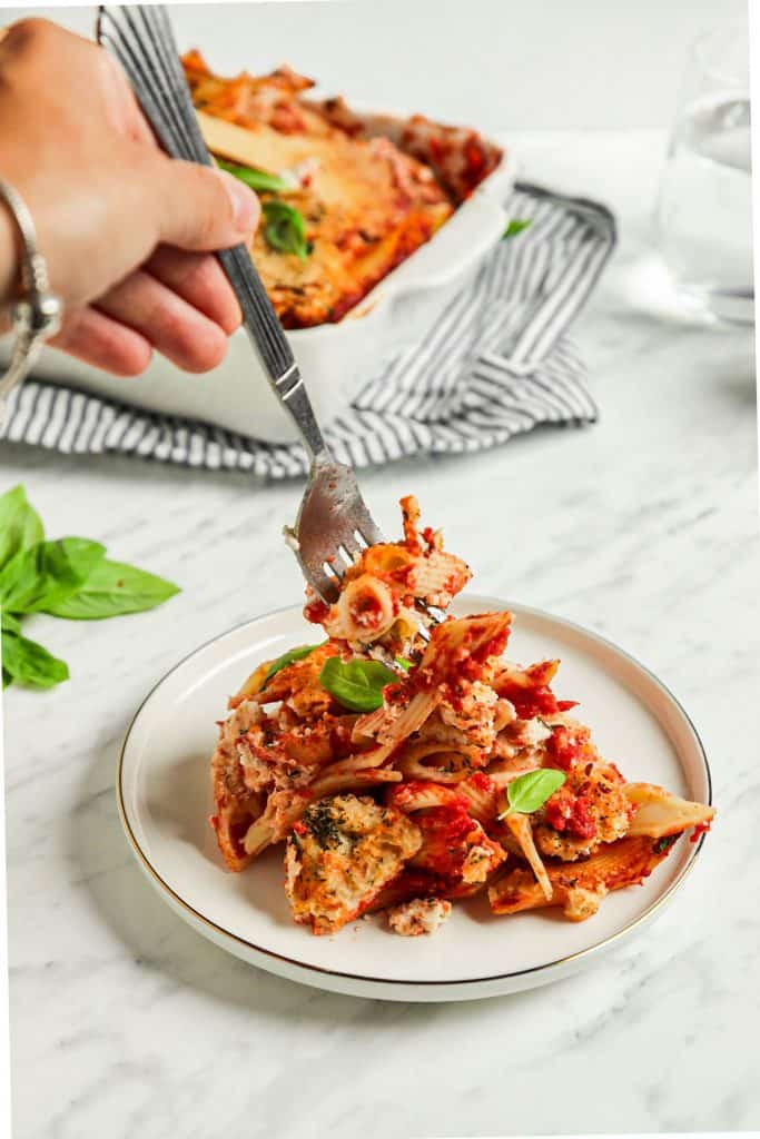 one fork digging into the vegan baked ziti on white plate in front of baking dish in background