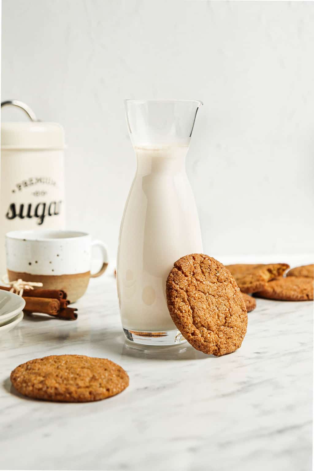 carafe of milk with pumpkin snickerdoodle on the side. pumpkin snickerdoodles in the background with mug, cinnamon, and sugar. 