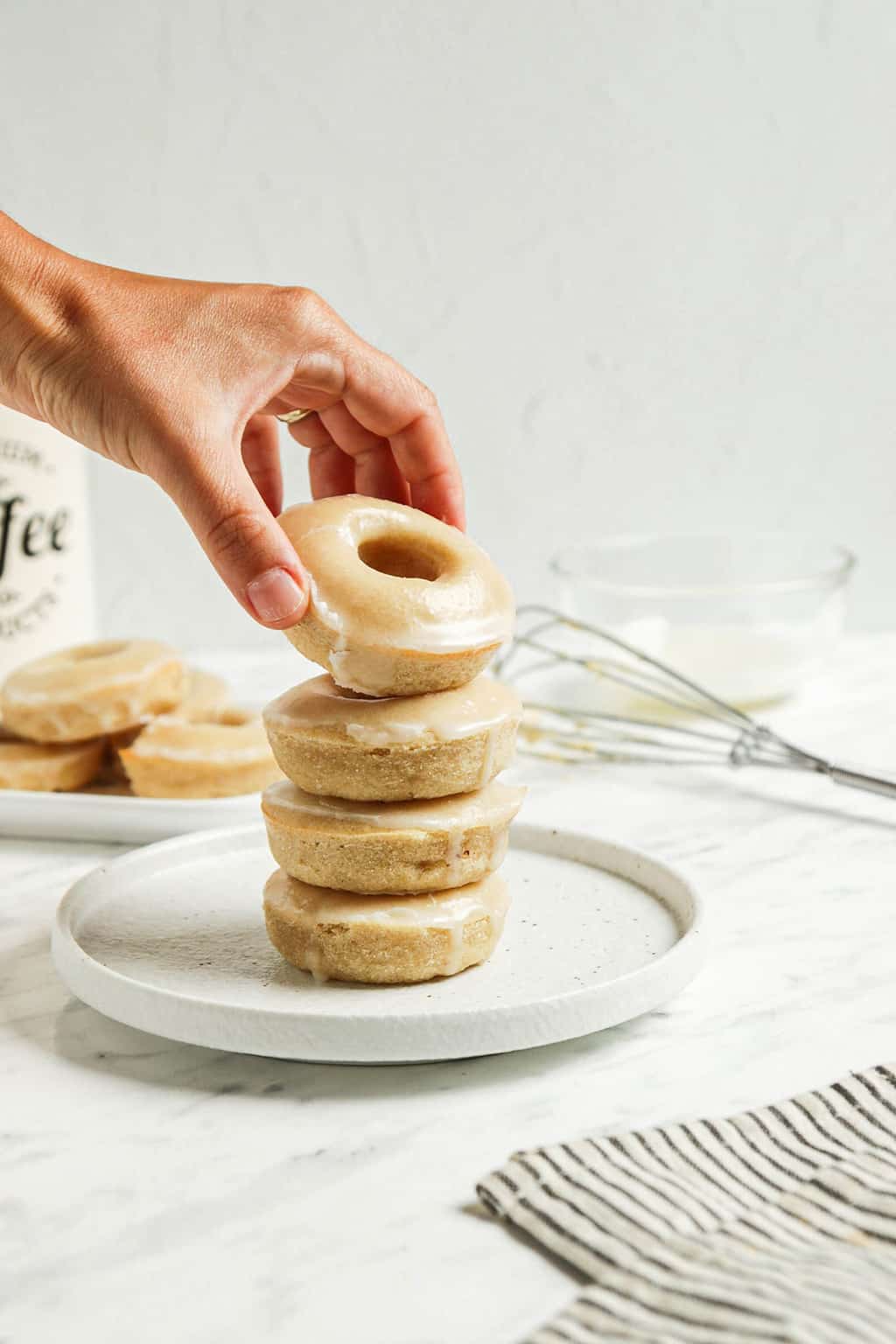 one stack of four vegan glazed doughnuts on white plate with vanilla icing and hand. One group of donuts and a whisk in the background