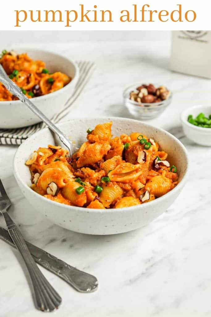 pumpkin alfredo with pasta in white bowl with utensils and green onions and hazelnuts in background