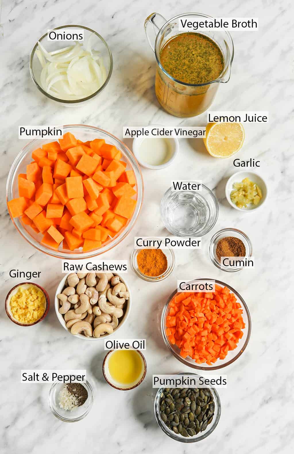 fresh pumpkin cubes, water, garlic, lemon juice, apple cider vinegar, onions, vegetable broth, cumin, curry powder, cashews, ginger, carrots, olive oil, salt, pepper, pumpkin seeds on white surface 