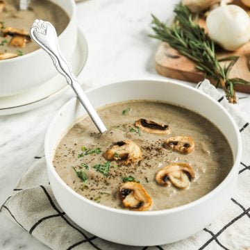 two bowls filled with creamy mushroom soup topped with whole mushrooms, salt, pepper, and fresh herbs with spoon