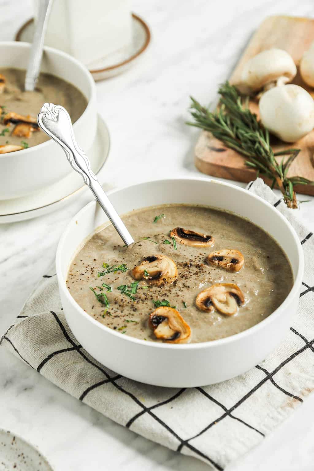 one bowl of creamy vegan mushroom soup with spoon and topped with whole mushrooms, salt, pepper, and chopped herbs