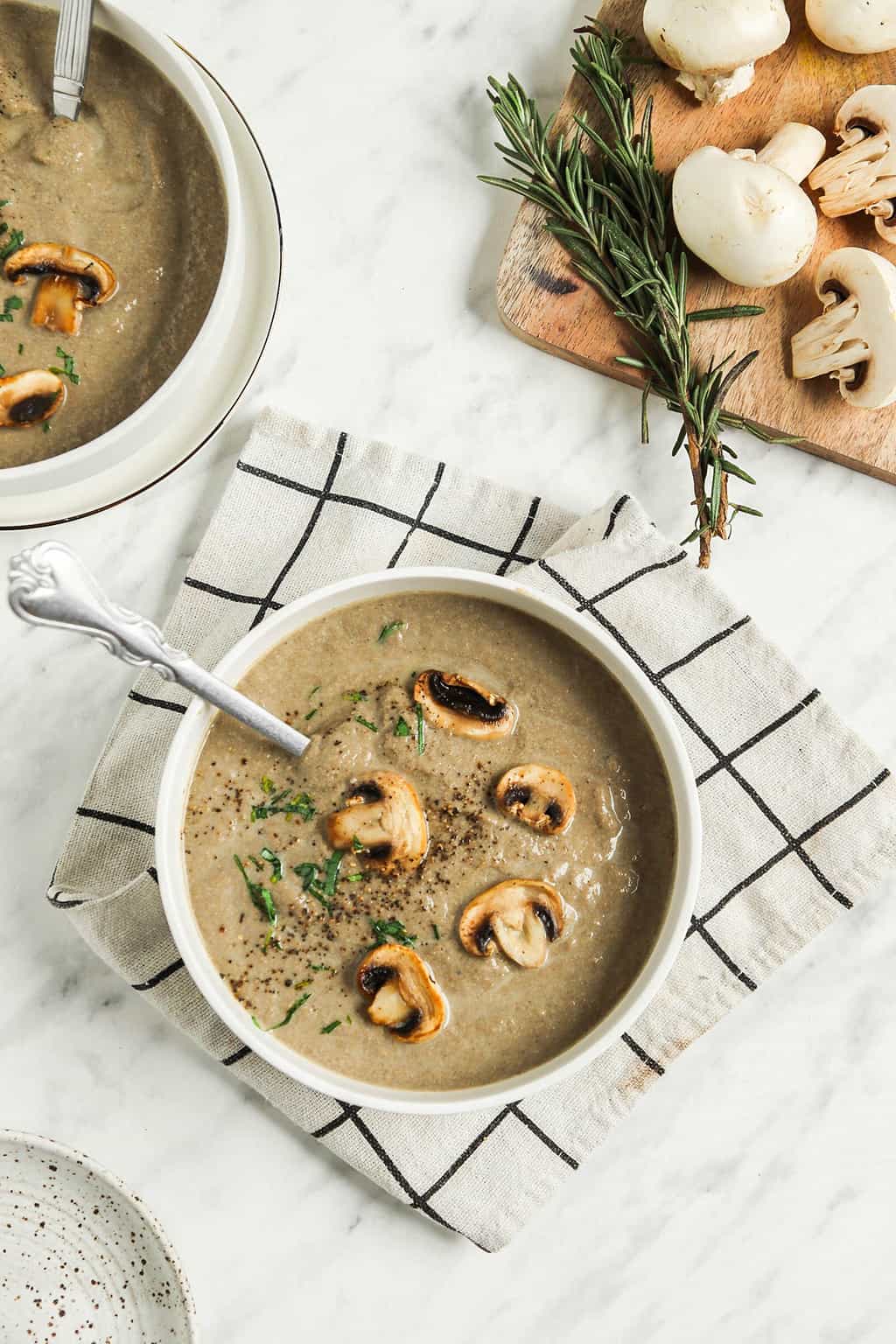 two bowls of vegan mushroom creamy soup with whole mushrooms, salt, pepper, and fresh herbs on top. Served with a spoon and checked linens. 