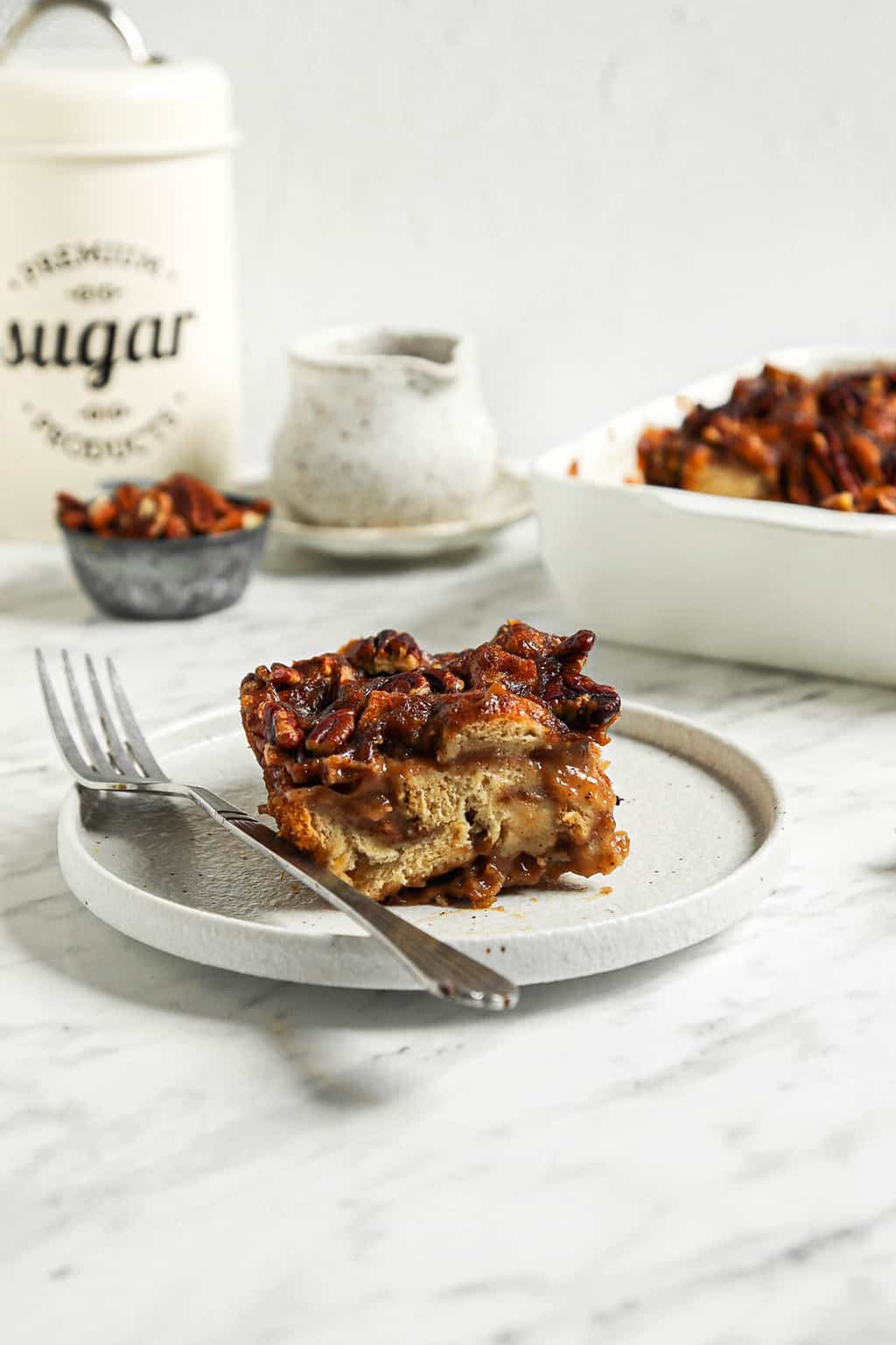 one slice of baked pecan bread pudding on white plate with fork and full baking dish in the background