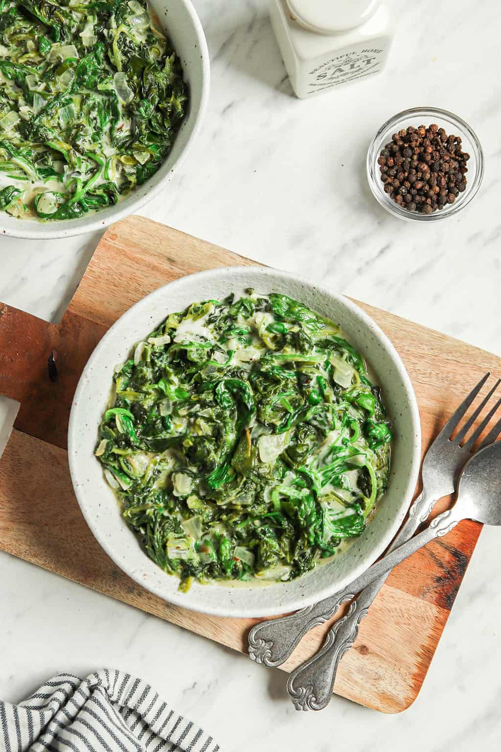 one white bowl filled with vegan creamed spinach with vegan cream and milk. Served with fork and spoon on wooden platter