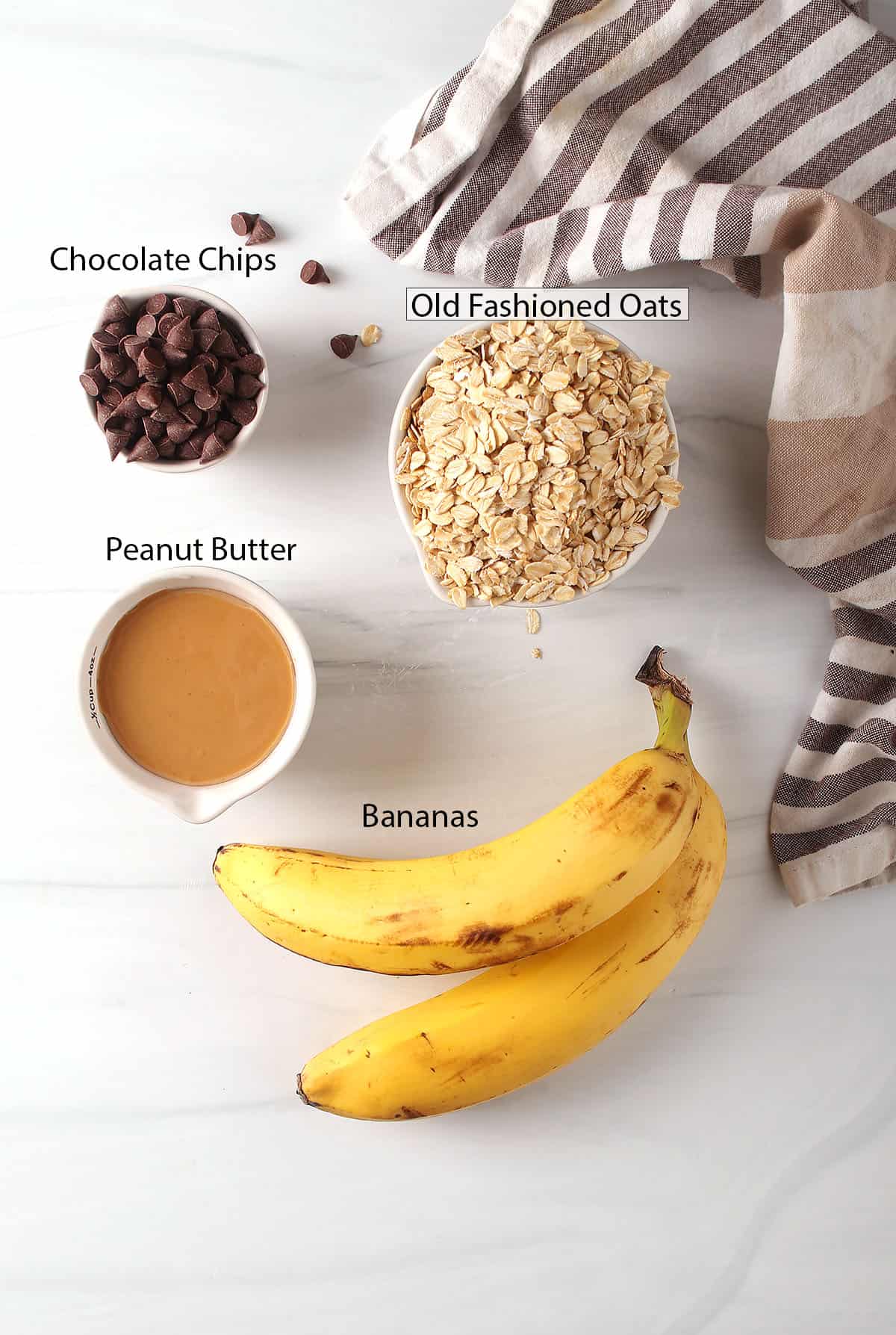 Peanut butter, chocolate chips, bananas, and oats measured out on a white marble countertop