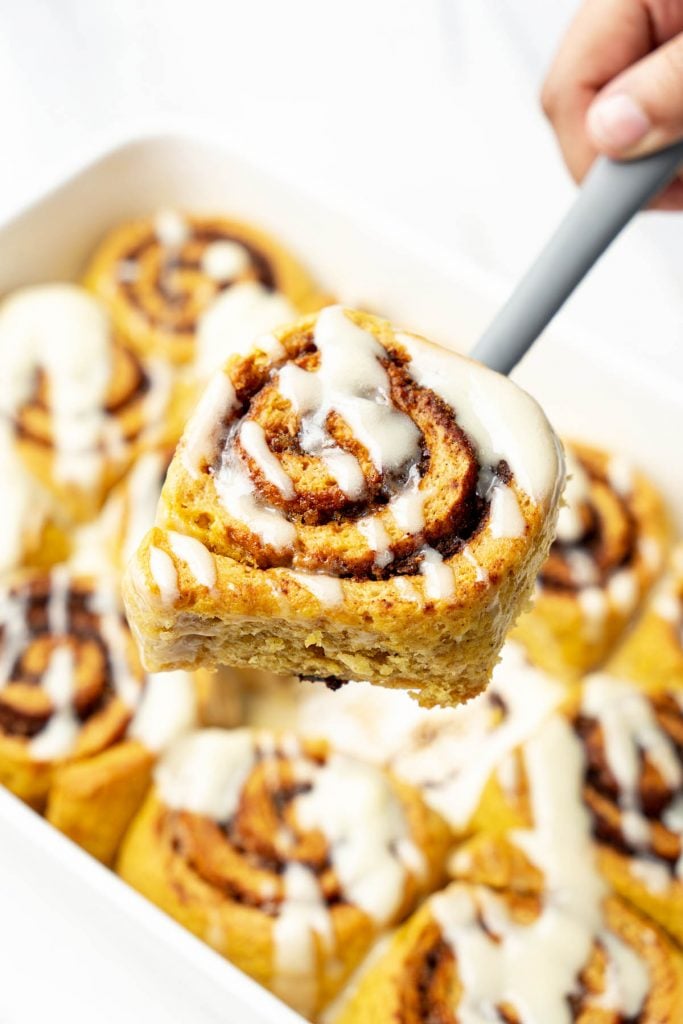 hand lifting a warm vegan pumpkin cinnamon roll with dairy-free maple glaze from a baking dish