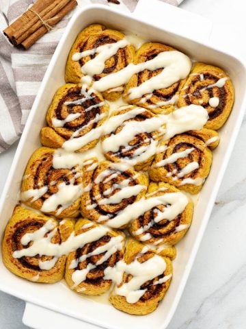 top-down view of warm vegan pumpkin cinnamon rolls in a white baking dish drizzled with creamy maple glaze