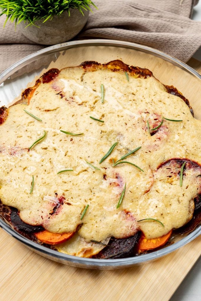 finished vegan vegetable gratin in a glass dish topped with cashew cream and fresh rosemary