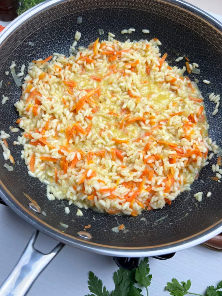 Carrot and arborio rice cooking in a skillet while absorbing broth during the process