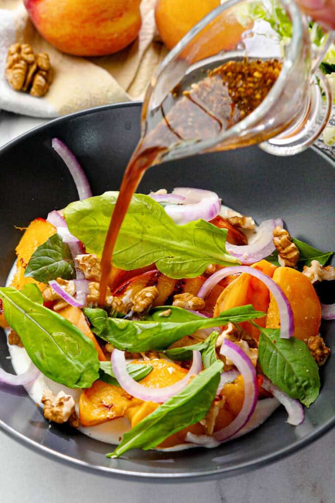 balsamic dressing being poured over a grilled peach salad with vegan feta, walnuts and arugula