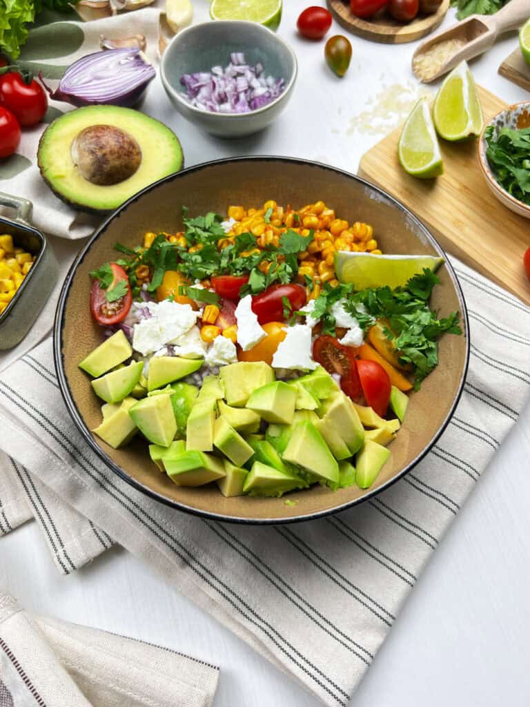 Vegan Mexican street corn bowl surrounded by avocado, lime, tomatoes and cilantro