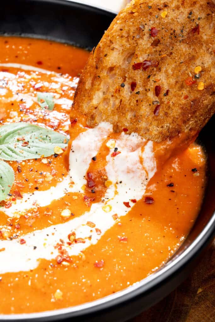 slice of toasted sourdough dipped into creamy roasted red pepper soup topped fresh herbs and seasonings