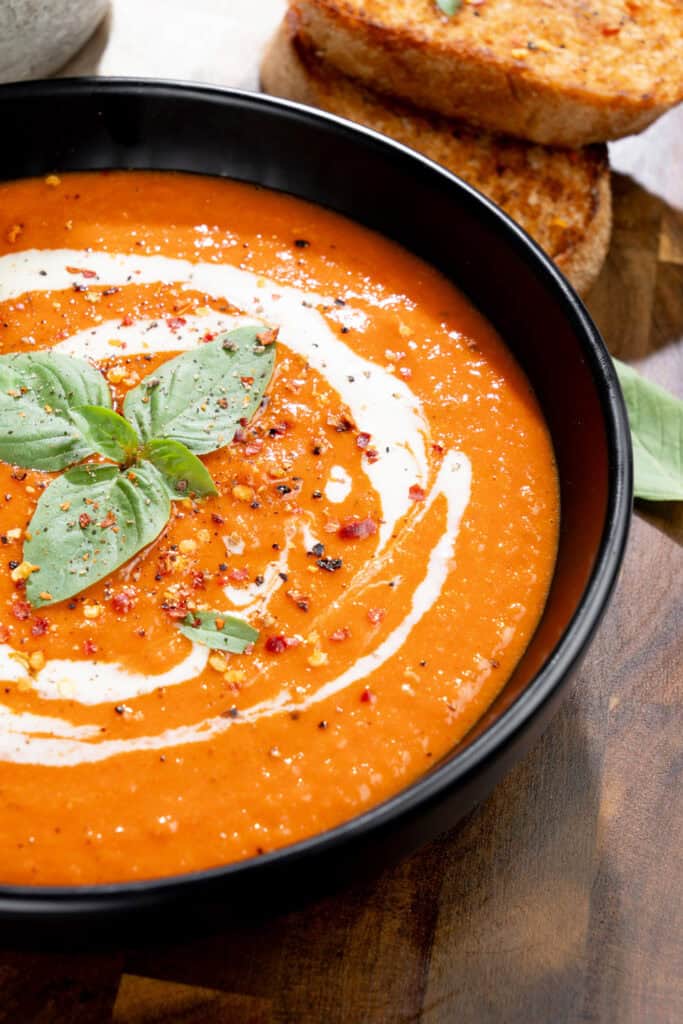 close-up of creamy roasted red pepper soup topped with a swirl of vegan cream, basil, oil and pepper